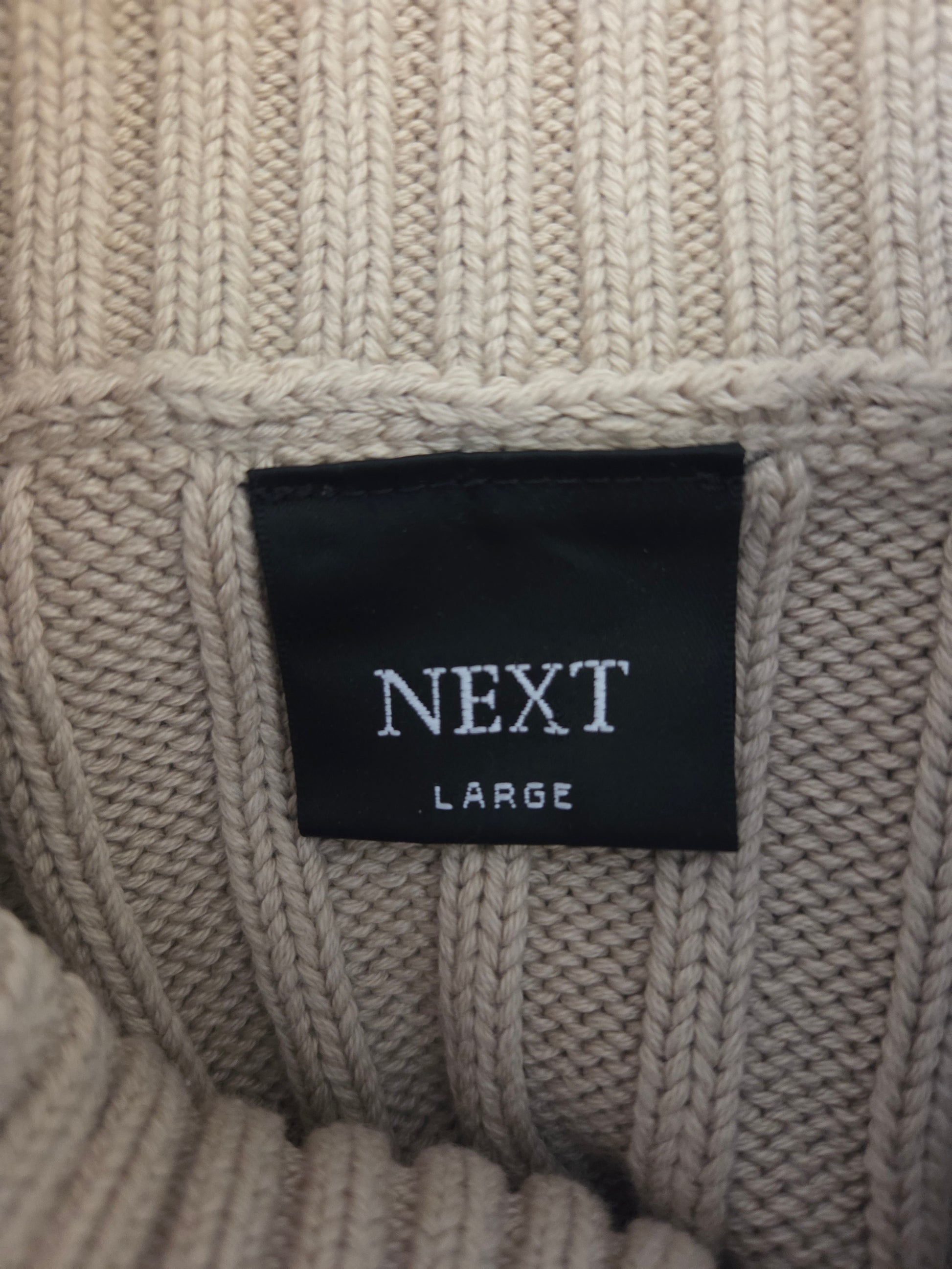 Close-up of a beige knit sweater with a 'Next' label on a textured fabric background