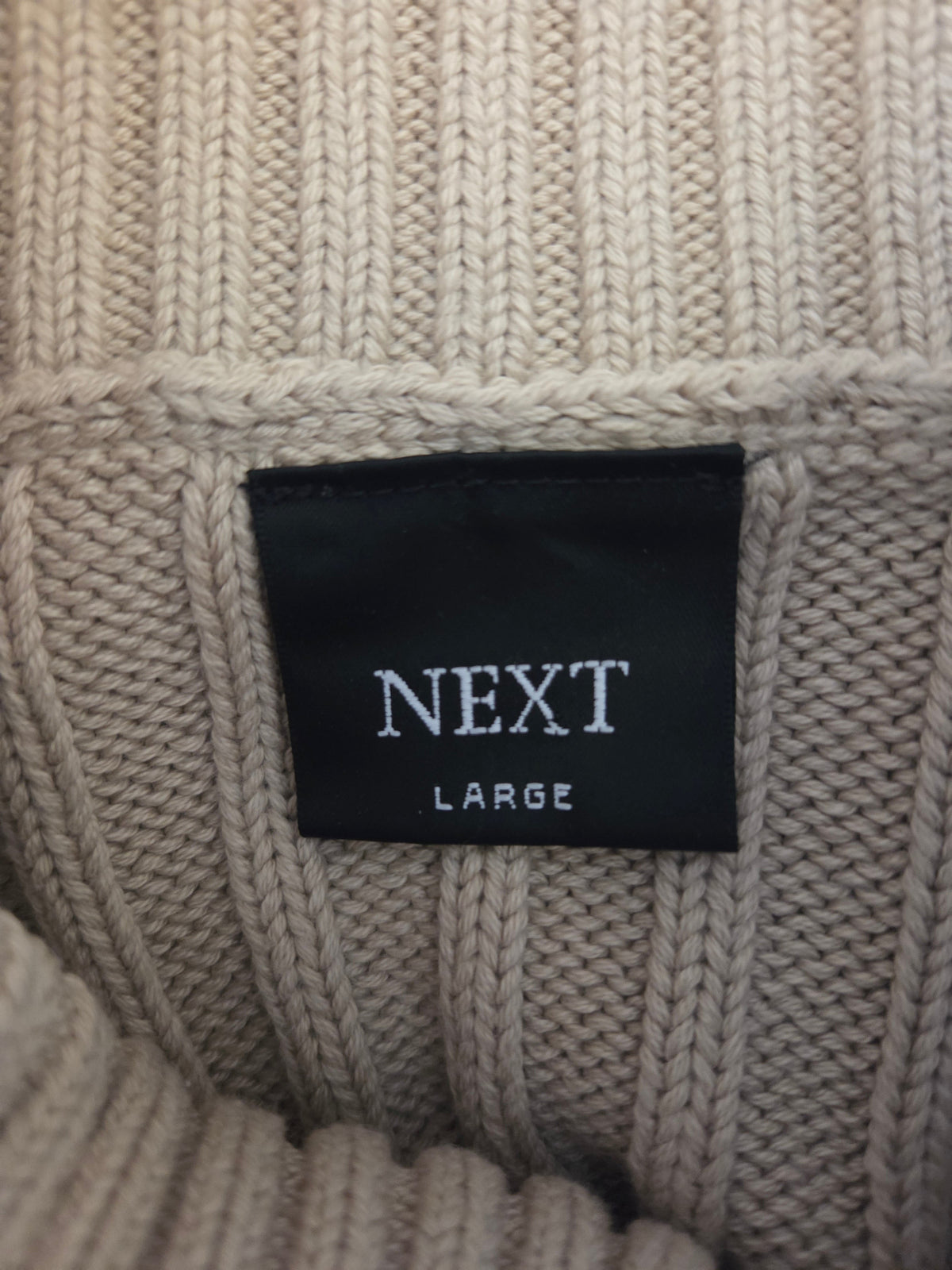 Close-up of a beige knit sweater with a 'Next' label on a textured fabric background