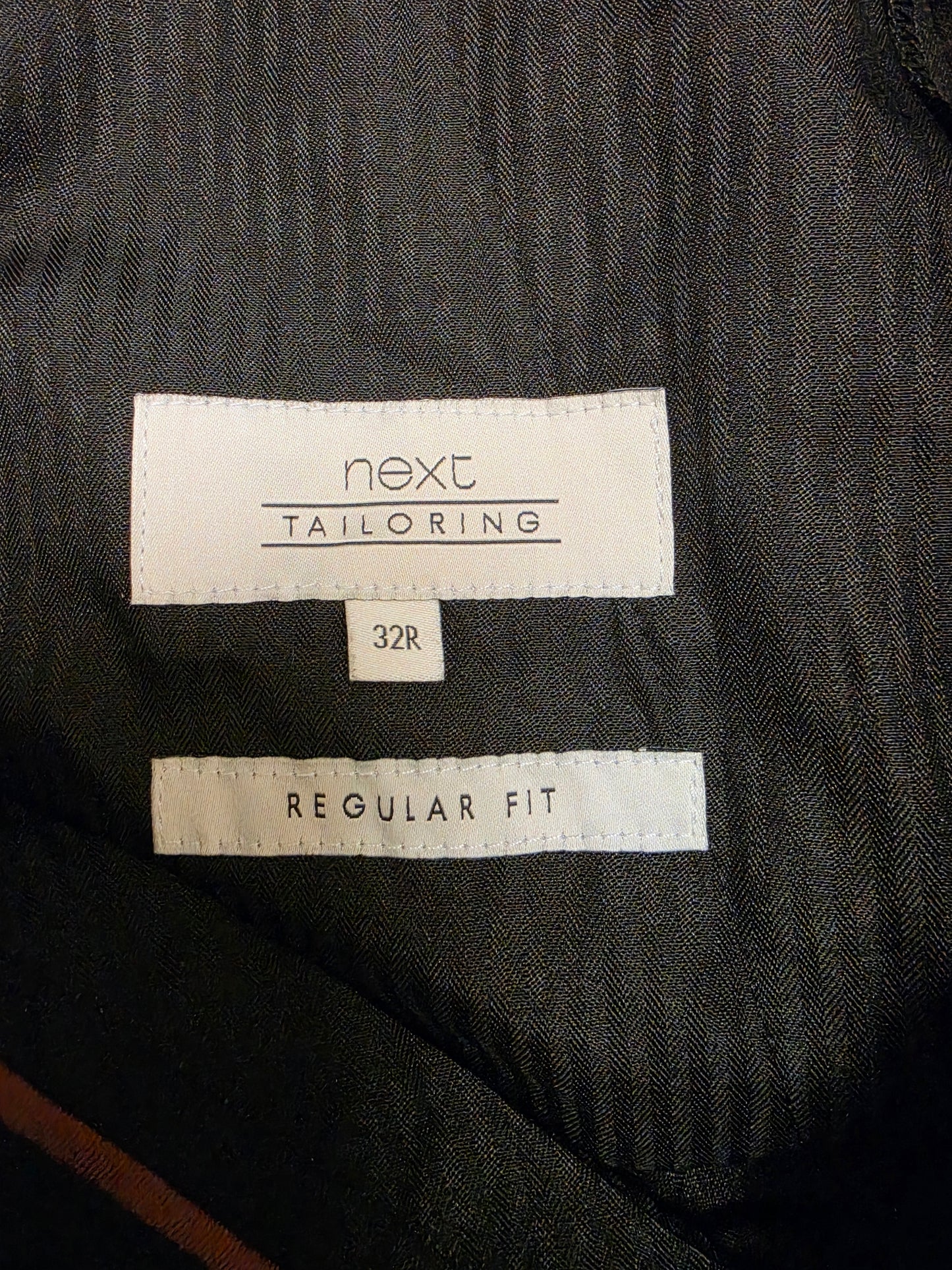 Close-up of a size label brand 'next TAILORING', size '32R', and 'REGULAR FIT' on a dark charcoal gray textured fabric.