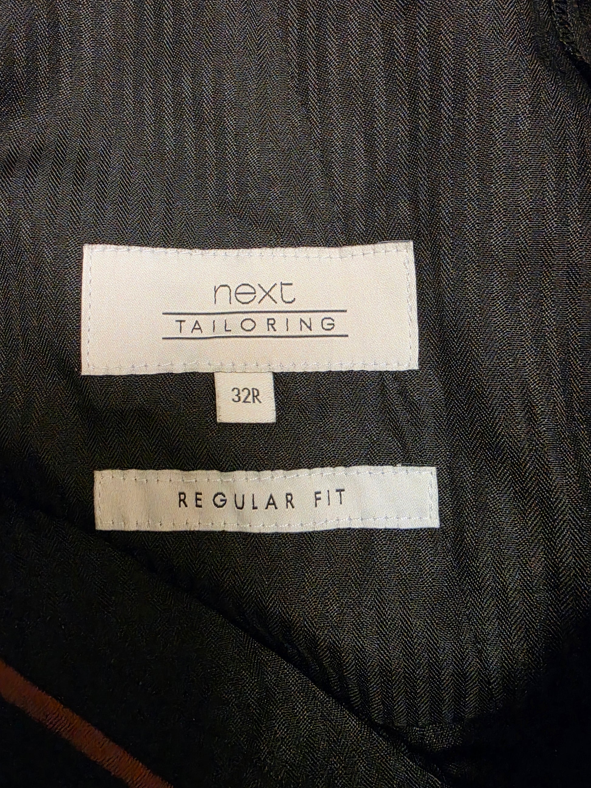 Close-up of a size label brand 'next TAILORING', size '32R', and 'REGULAR FIT' on a dark charcoal gray textured fabric.